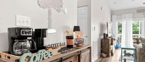 A coffee bar area with a coffee machine, a sign reading 'Coffee Bar,' and a coffee cup. There is a wooden cabinet with drawers and a small table with a lamp.