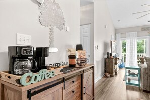 A coffee bar area with a coffee machine, a sign reading 'Coffee Bar,' and a coffee cup. There is a wooden cabinet with drawers and a small table with a lamp. - 2 bed 2 bath condo in Ocean Keyes, N. Myrtle Beach (North Myrtle Beach)