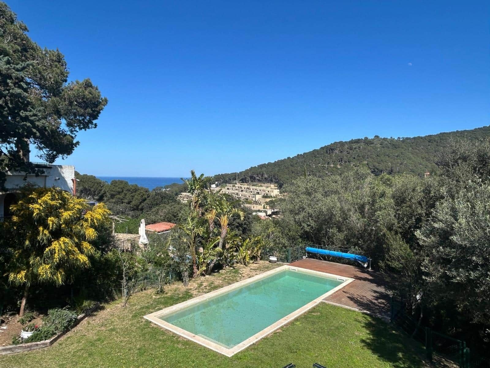 A pool surrounded by a garden with a view of a coastal town and hills.