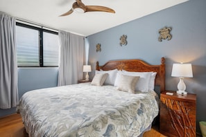 A hotel room with a large bed, two bedside tables, a ceiling fan, and a window with curtains. - Polynesian Shores 114 by Gather - Oceanfront 1BR w/ Pool & Ocean Views (Lahaina)
