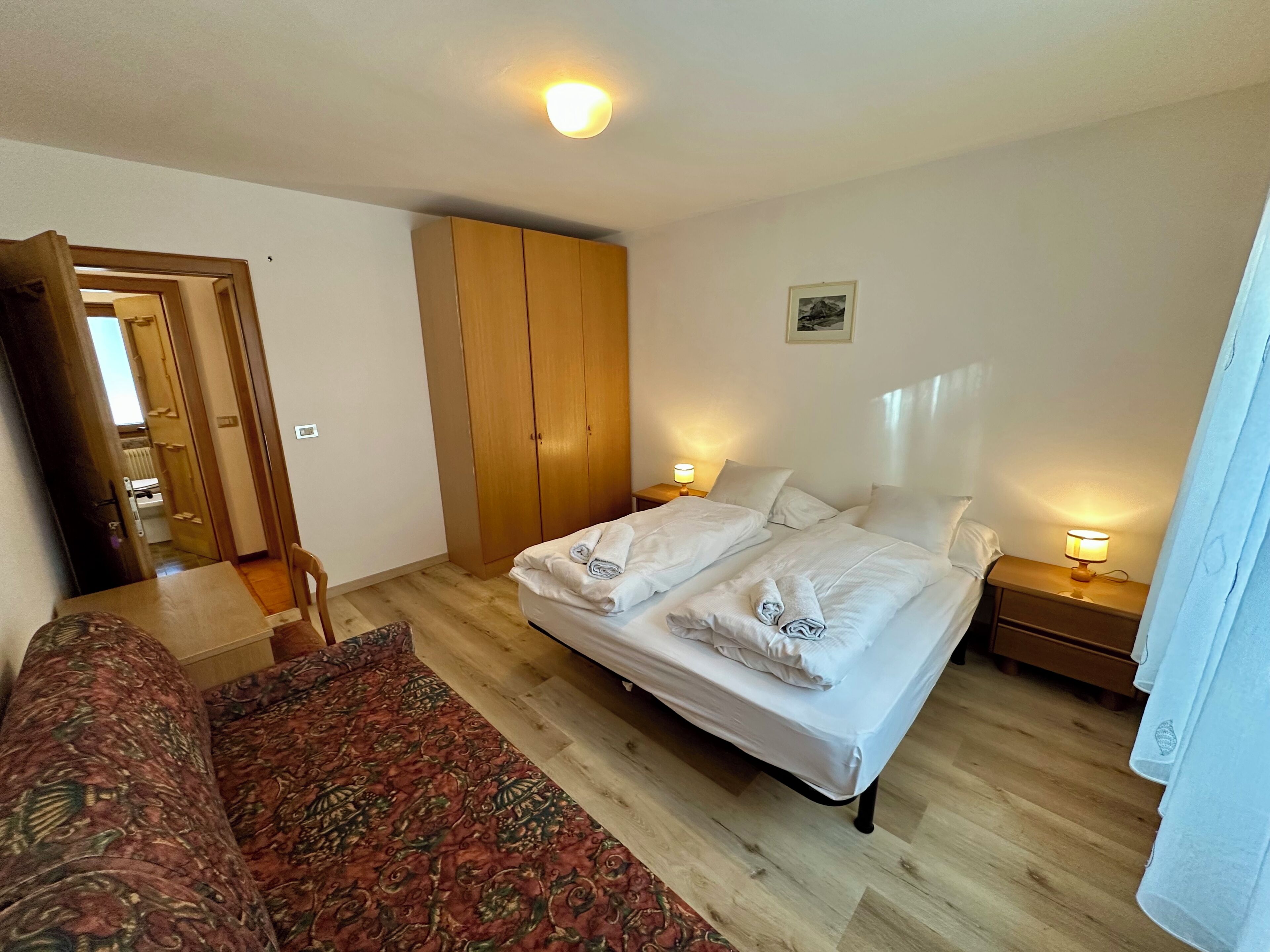 Ein Schlafzimmer mit einem Doppelbett, einem Holzschrank, einem Nachttisch mit Lampe und einem gemusterten Sofa.