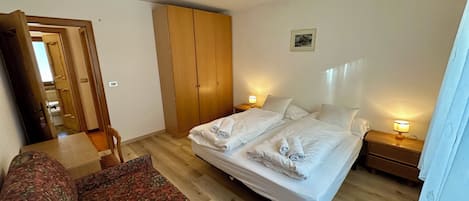 A bedroom with a double bed, a wooden wardrobe, a bedside table with a lamp, and a patterned sofa.