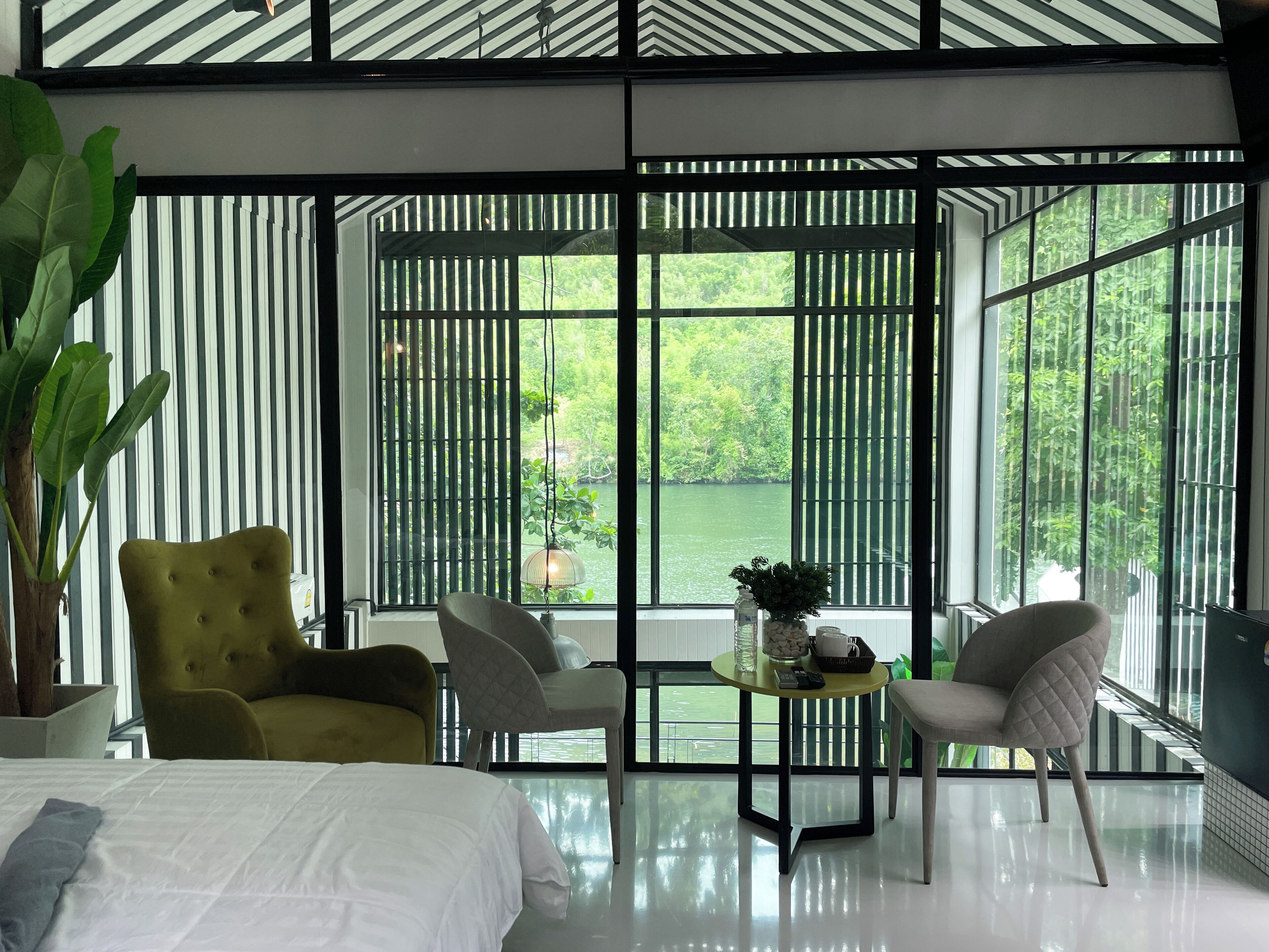 Une chambre d’hôtel moderne avec un grand lit, deux fauteuils, une petite table avec un vase, et une vue sur une rivière grâce à de grandes fenêtres équipées de stores verticaux.