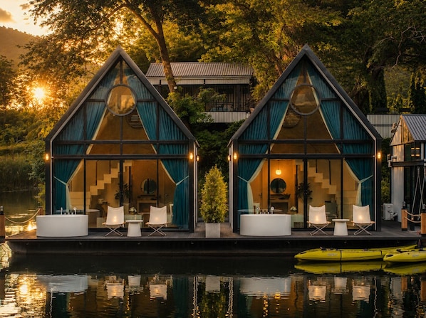 A modern houseboat with a glass facade, outdoor seating, and a kayak nearby.