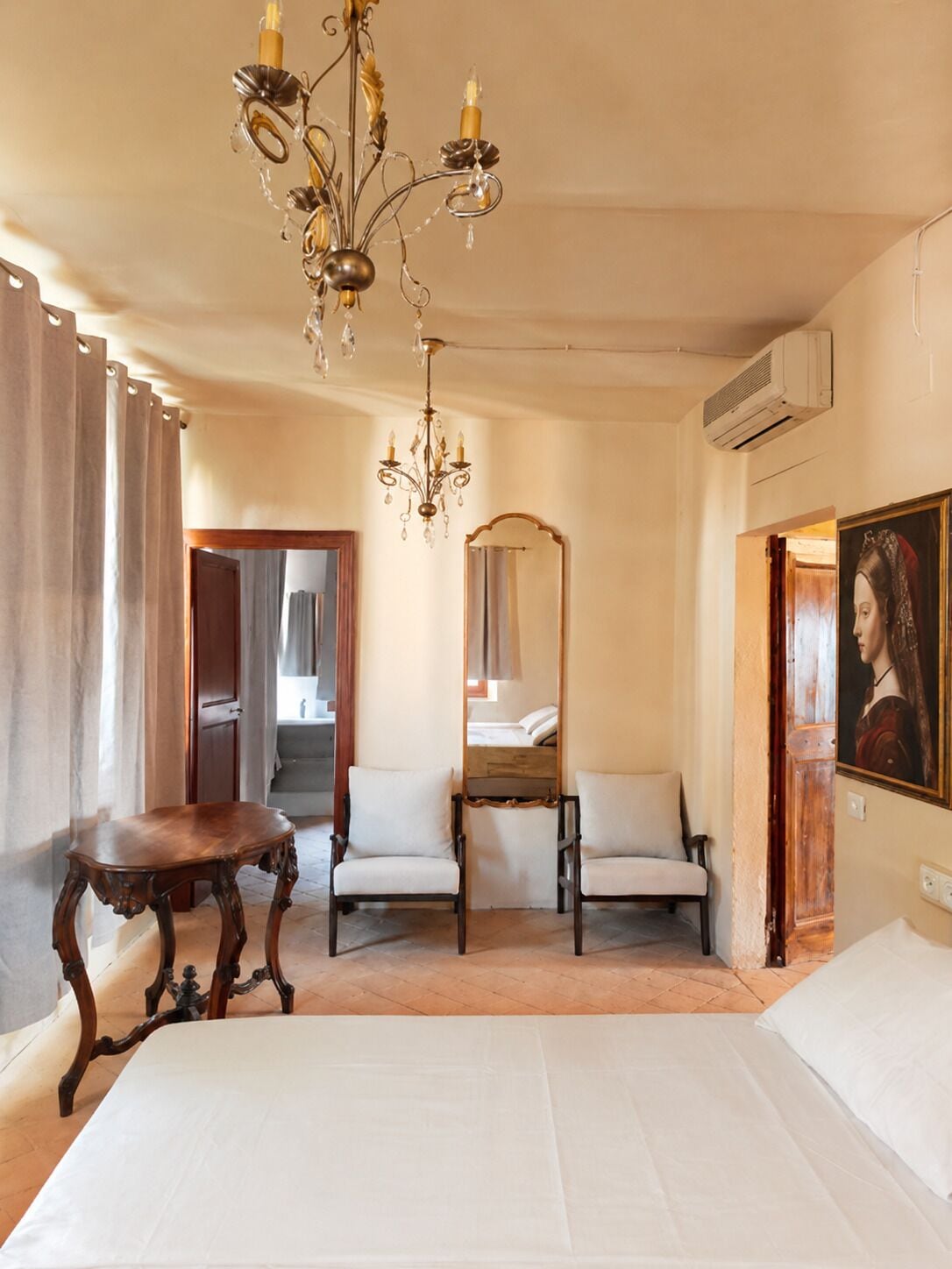 A hotel room with a bed, two chairs, a table, a chandelier, a mirror, and a painting.