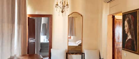 A hotel room with a bed, two chairs, a table, a chandelier, a mirror, and a painting.