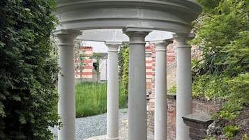 Ein Pavillon mit Metallveranda und weißen Säulen, umgeben von Grünpflanzen und einer Backsteinmauer.