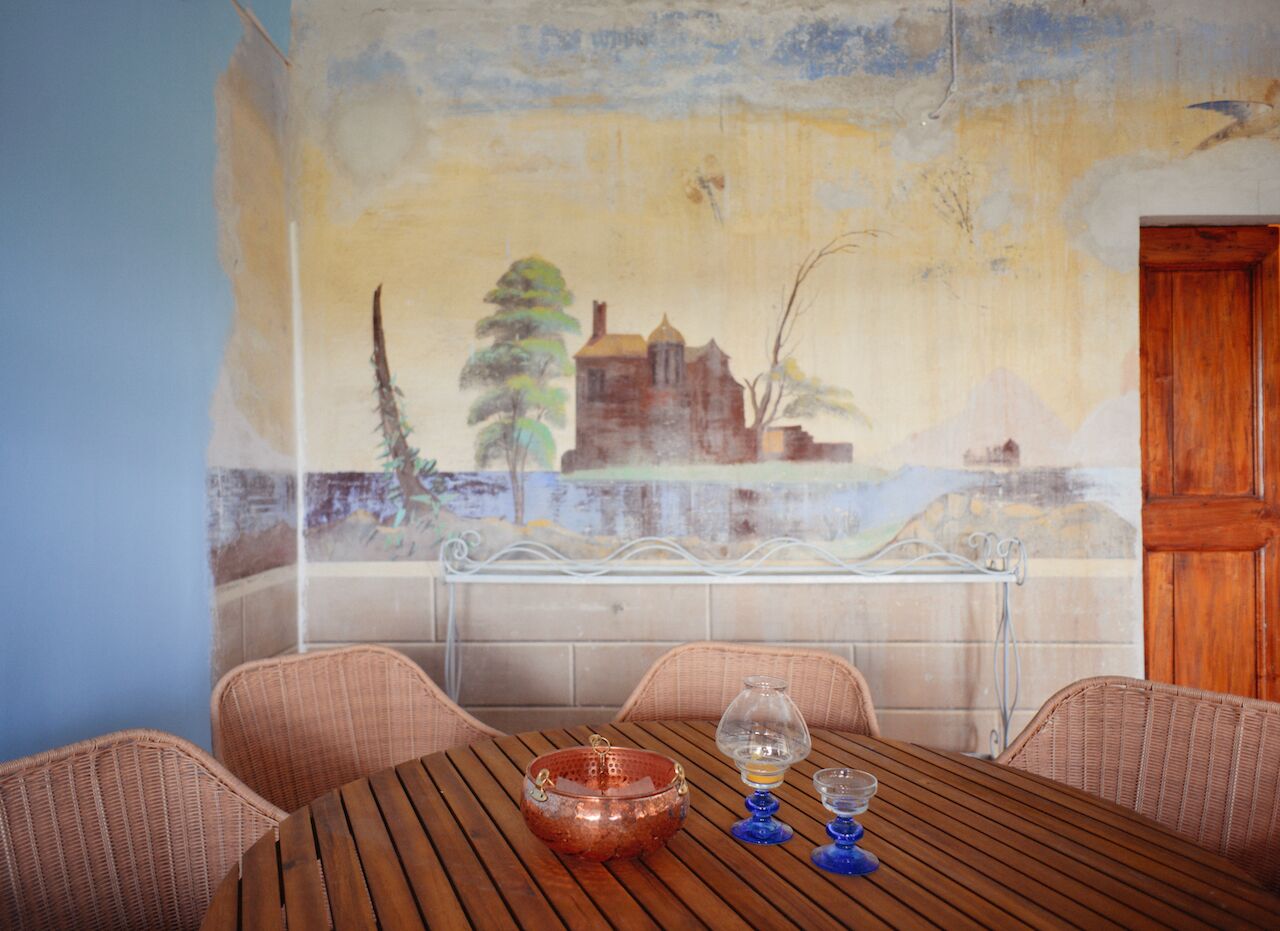 Un coin repas avec une table dressée pour deux personnes, un banc en métal, des chaises en osier et une fresque murale.