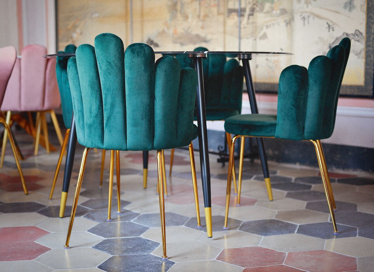 Un'area pranzo con sedie rivestite in tonalità petrolio e gambe dorate, un tavolo in vetro e un pavimento piastrellato.