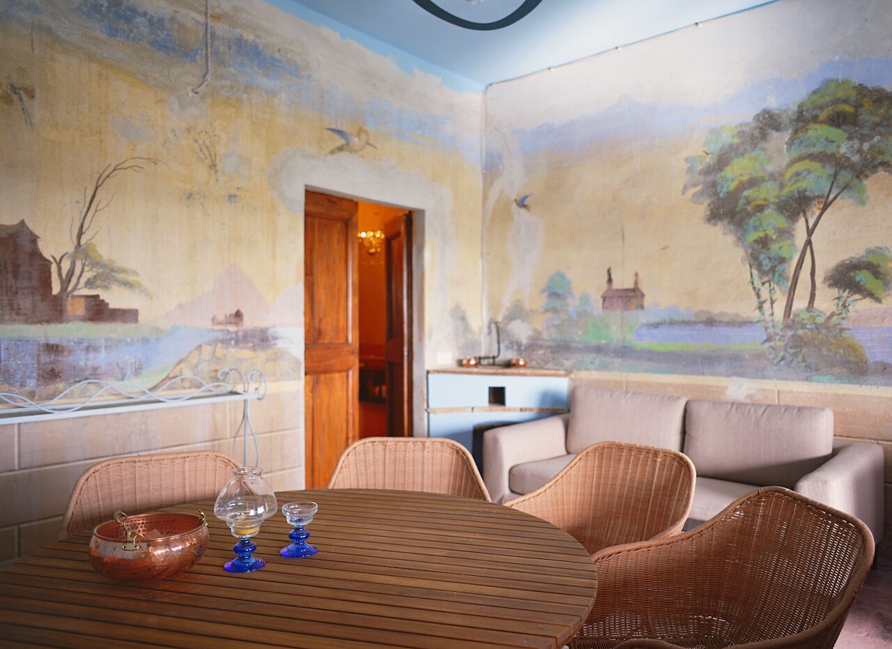 A dining area with a wooden table, wicker chairs, and a view of a landscape painting on the wall.