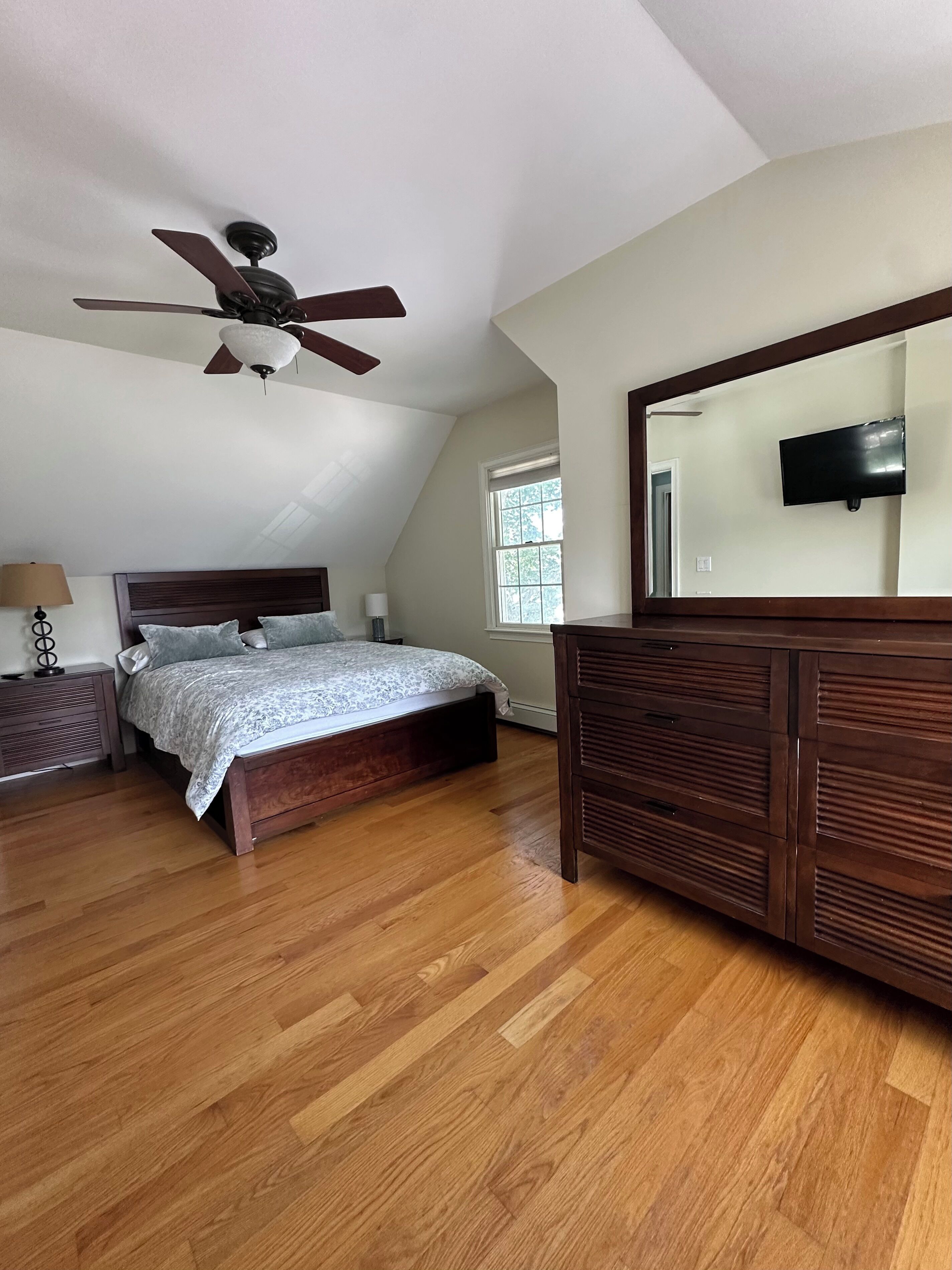 Une chambre à coucher comprenant un lit, une commode, un ventilateur de plafond, une fenêtre et un téléviseur fixé au mur.