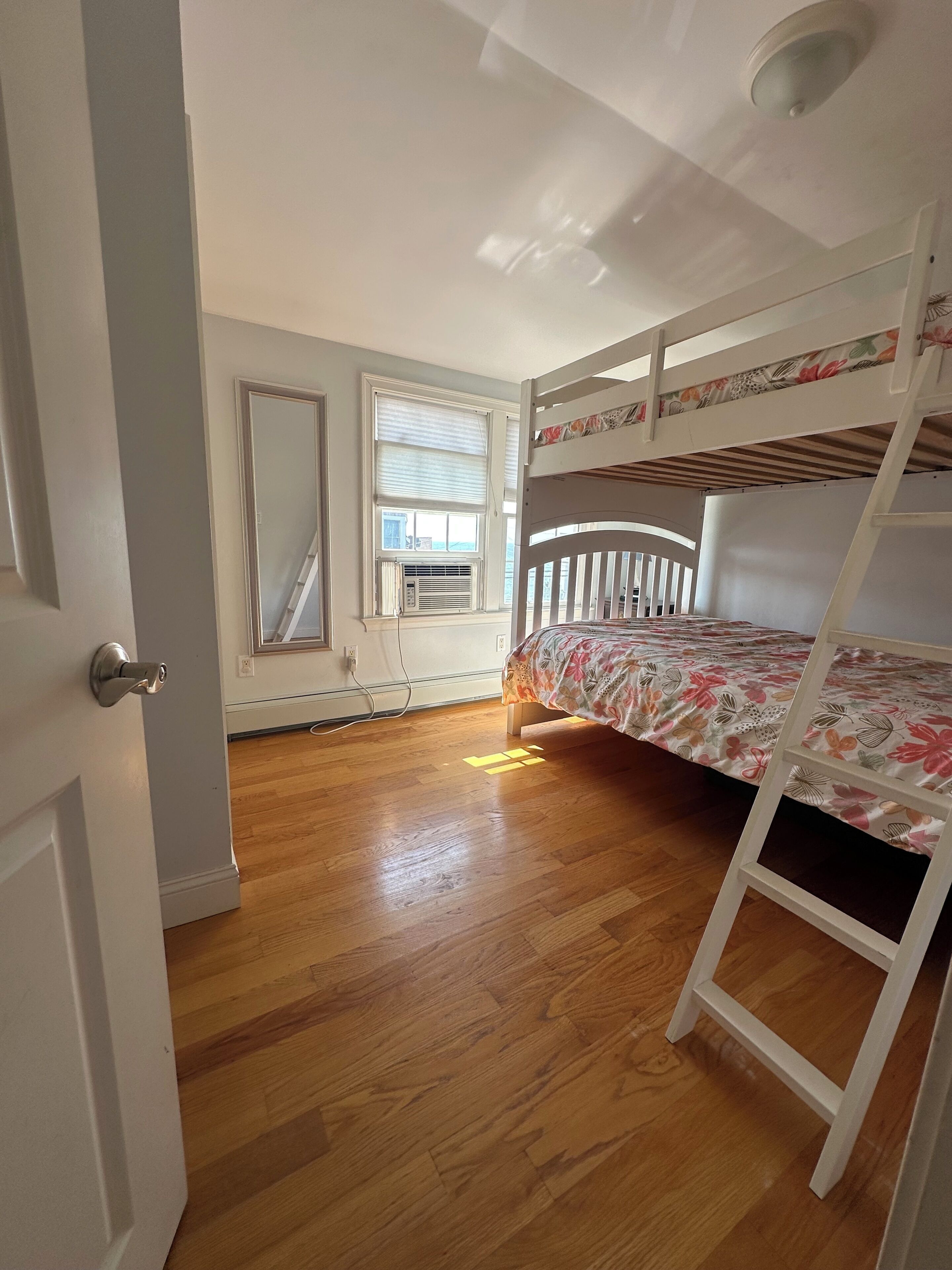 A room with a bunk bed, wooden flooring, a radiator, and a window with blinds.