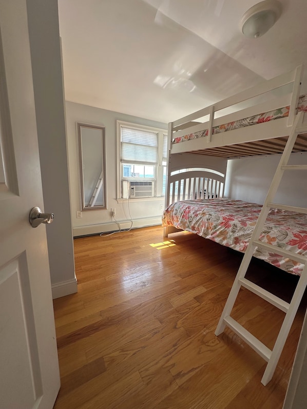 A room with a bunk bed, wooden flooring, a radiator, and a window with blinds.