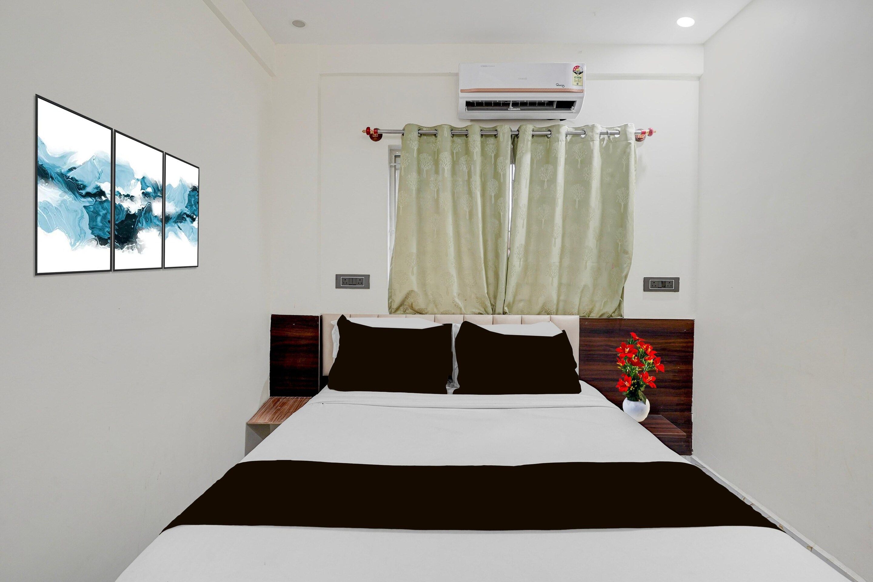A hotel room with a bed, a nightstand, a wall-mounted air conditioner, a window with curtains, and a framed abstract painting.