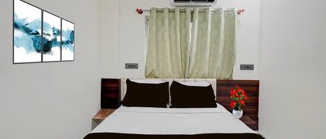 A hotel room with a bed, a nightstand, a wall-mounted air conditioner, a window with curtains, and a framed abstract painting.