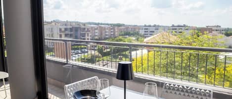 A balcony with a glass table, chairs, and a view of buildings and greenery.