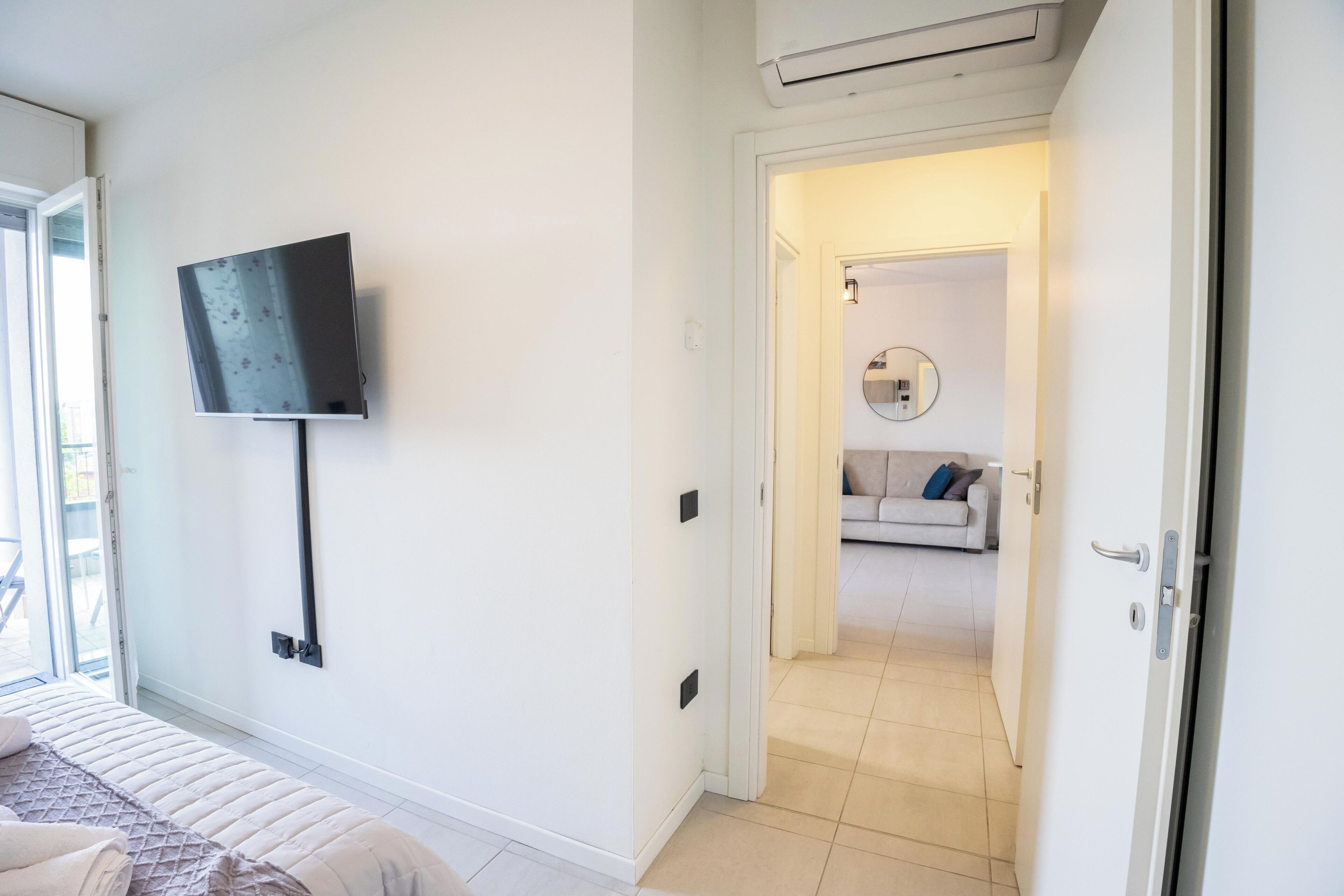 Ein Hotelzimmer mit einem an der Wand befestigten Flachbildfernseher, einem Bett mit gesteppter Bettdecke und einer Tür zu einem Wohnbereich mit Sofa und rundem Spiegel.