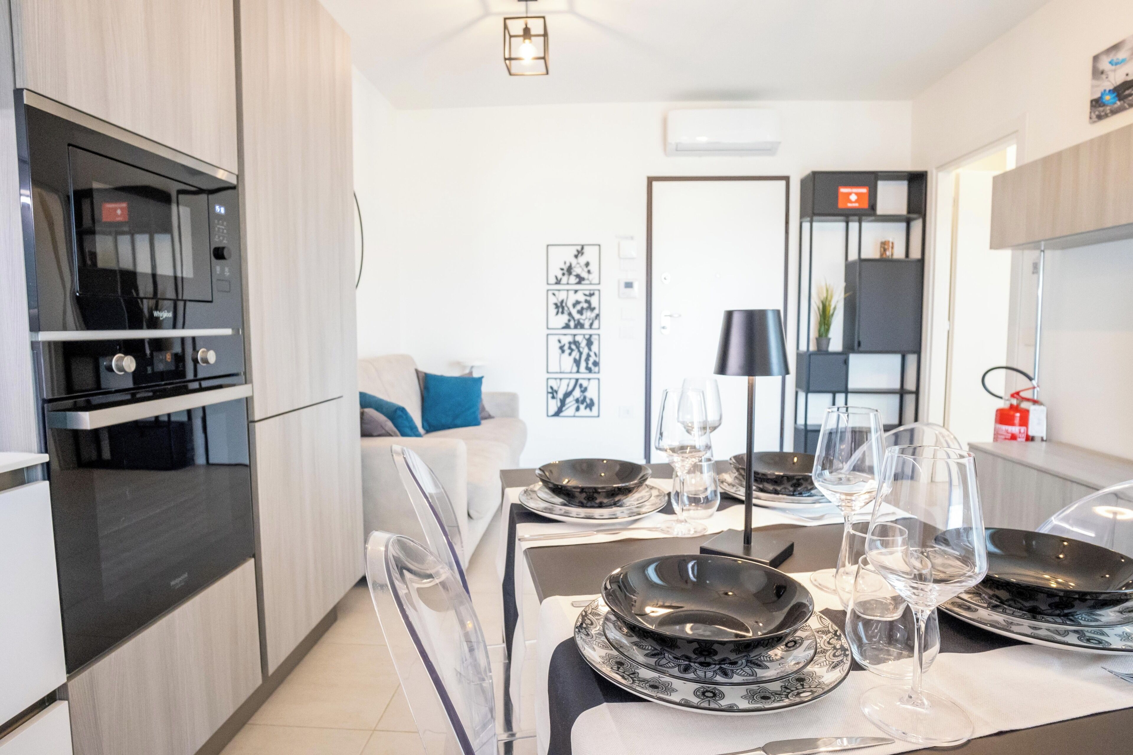 Una cocina moderna con horno y microondas integrados, una zona de comedor con una mesa dispuesta para dos personas, y una sala de estar con un sofá y una mesa de centro.