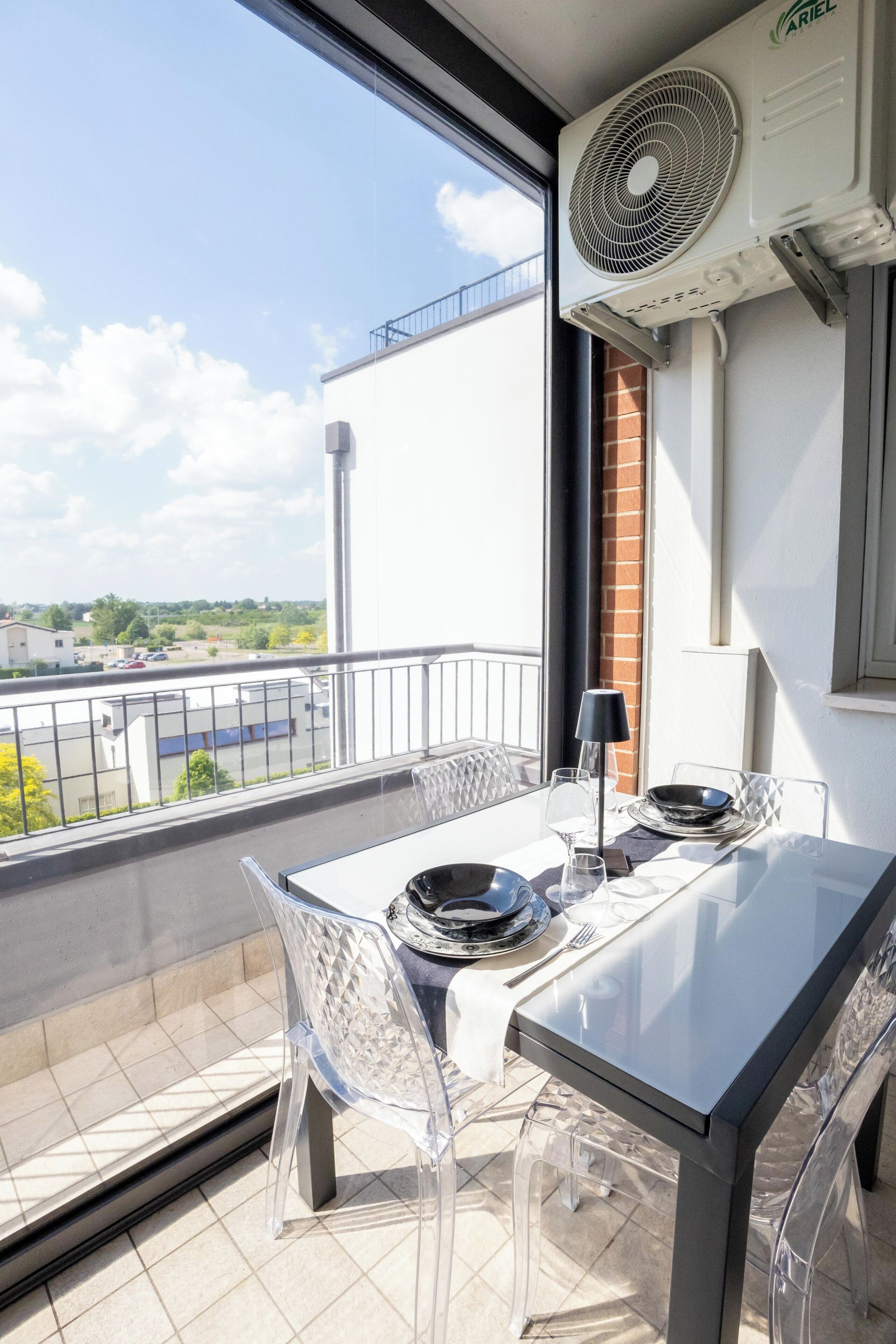 Un balcone con un tavolo apparecchiato per mangiare, sedie trasparenti, una ventola e vista sulla città.