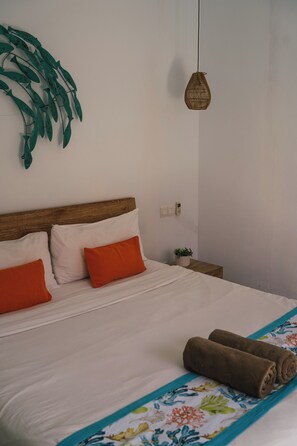 Ein Bett mit weißer Bettwäsche, zwei orangefarbene Kissen, ein braunes Handtuch und eine gewebte Leuchte.