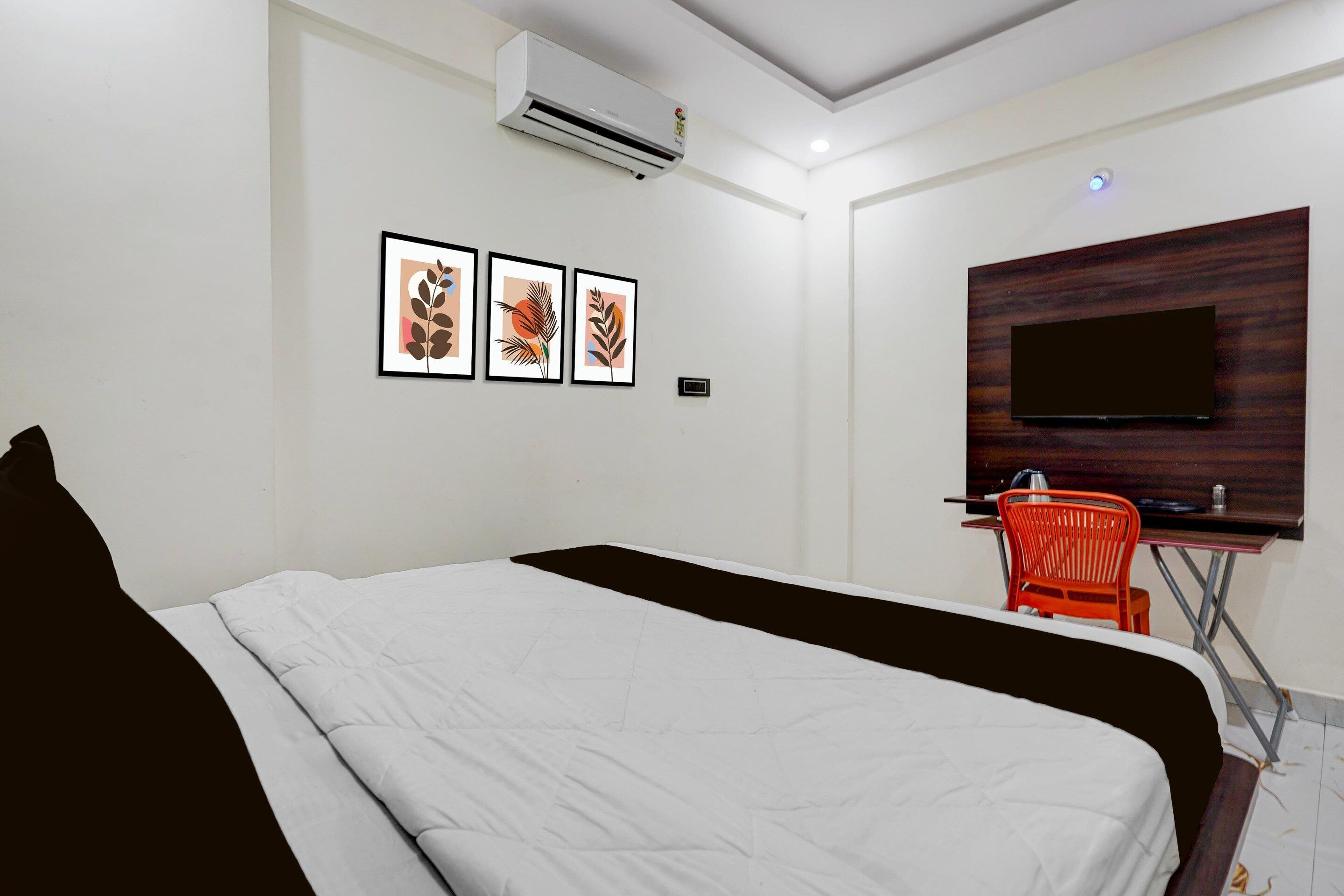 A hotel room with a bed, a desk with a chair, a flat-screen TV, and three framed artworks on the wall.
