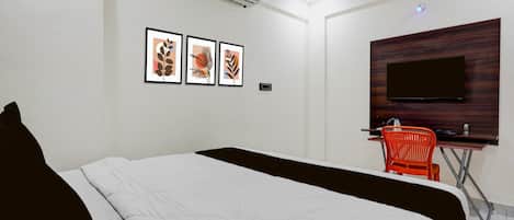 A hotel room with a bed, a desk with a chair, a flat-screen TV, and three framed artworks on the wall.