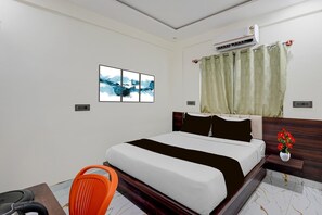 A hotel room with a bed, a chair, a nightstand, an air conditioner, a window with curtains, and a wall-mounted artwork.