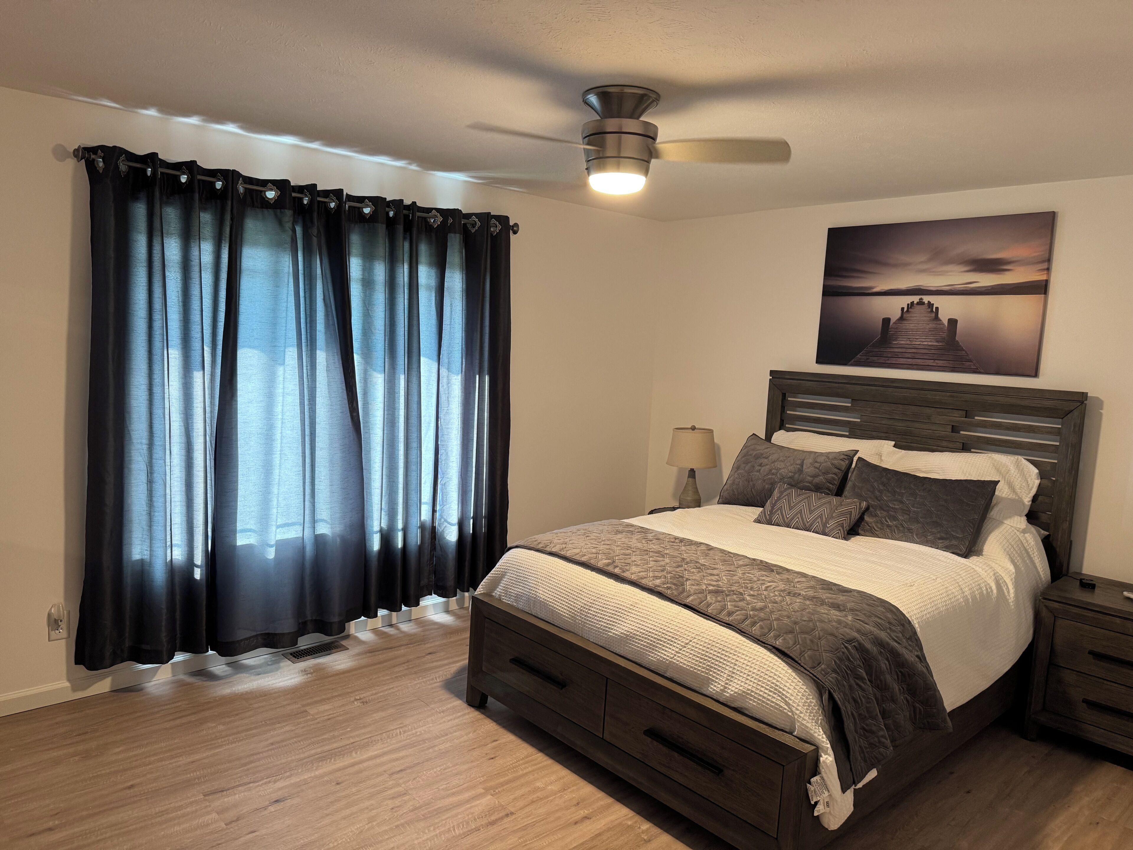 Un dormitorio con cama, mesita de noche, lámpara, ventilador de techo y un cuadro en la pared.