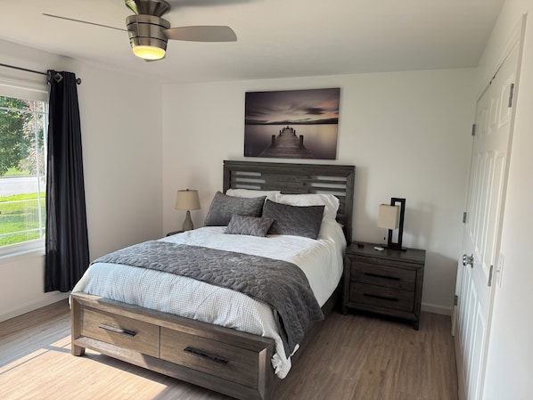 A bedroom with a bed, bedside tables, a ceiling fan, a window with curtains, and a wall-mounted artwork.