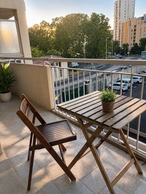 Un balcon avec une table et des chaises en bois, un pot de fleurs et une vue sur un parking et des immeubles.