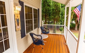 A covered porch with two chairs and a table, an American flag, and a framed picture on the wall.