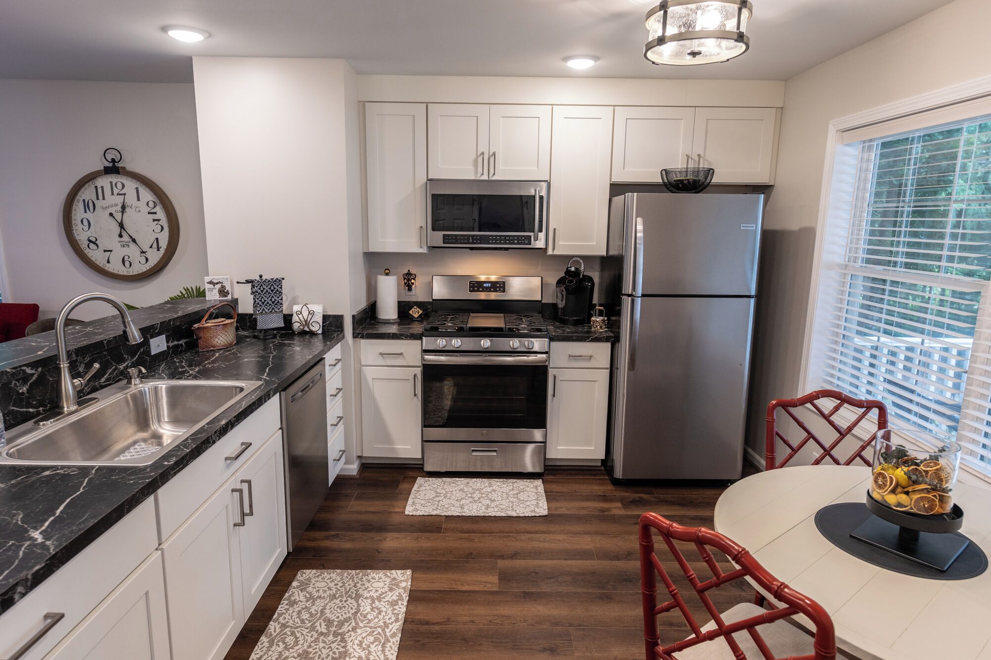 A modern kitchen with white cabinets, stainless steel appliances, a marble countertop, and a dining area with a red chair.