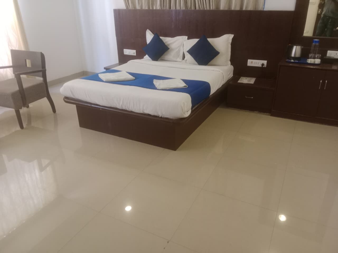 Habitación de hotel con una cama grande, cabecera de madera, mesita de noche con lámpara y una silla.