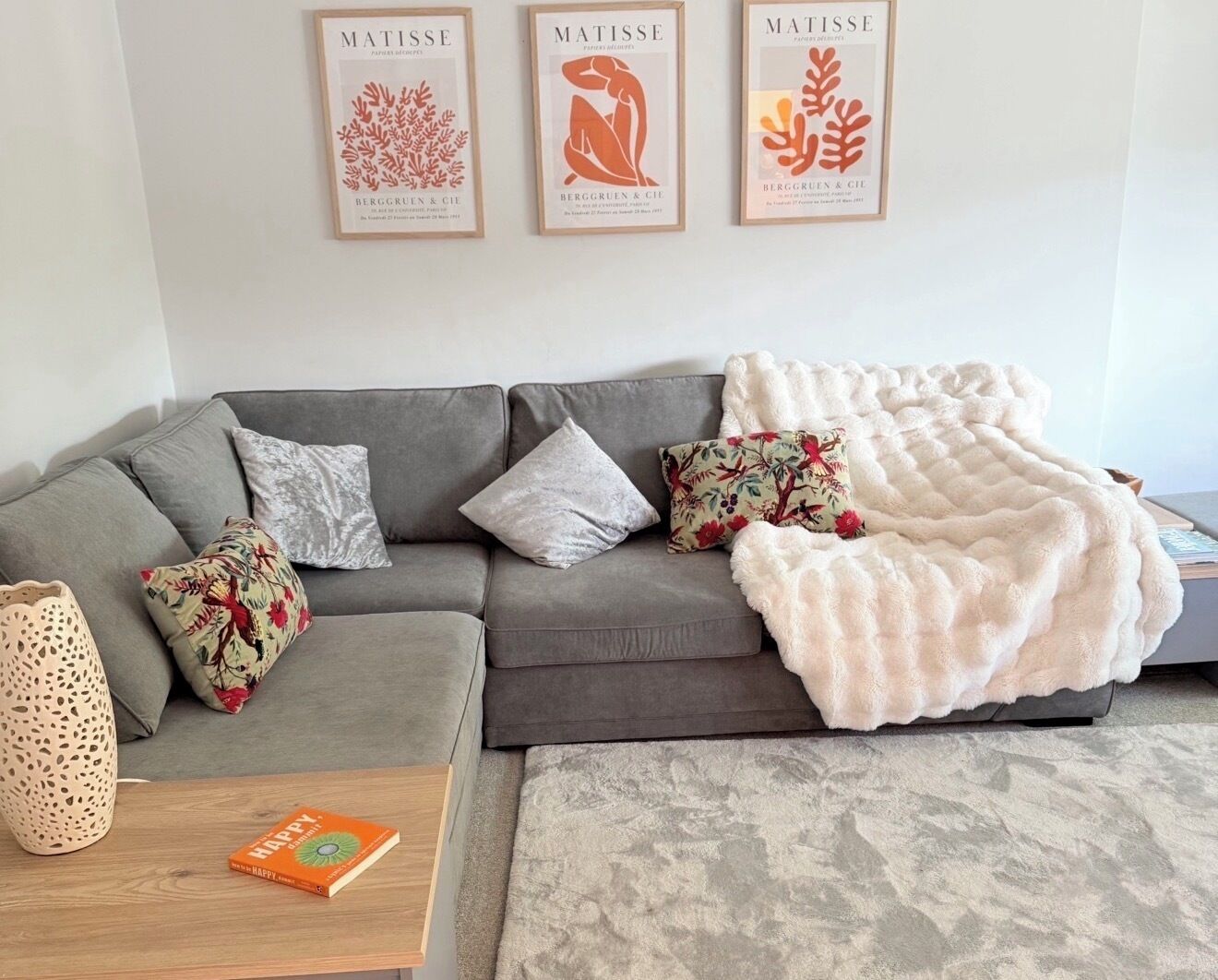 A modern living room with a grey sectional sofa, decorative pillows, a white furry throw blanket, and three framed artworks on the wall.
