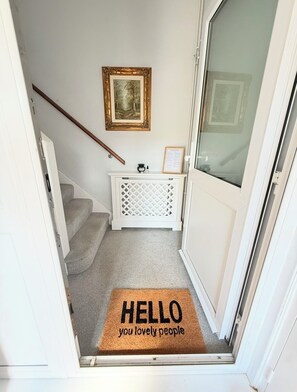 A hallway with a carpeted staircase, a white console table, a framed picture on the wall, and a doormat with 'HELLO you lovely people' text. - Nr Windsor & Legoland - Sleeps 5 - Free Parking (Maidenhead)