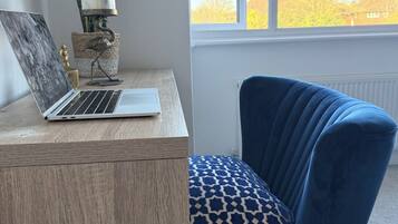 A room with a wooden desk, a laptop, a blue chair, a window with a view of trees, and a lamp.