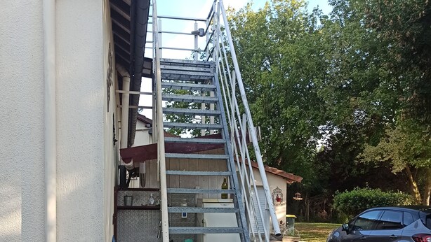A metal staircase leading to a building with a garage door, a parked car, and a potted plant. - Small studio on the banks of the Eure – quiet, nature & large outdoor spaces (Garennes-sur-Eure)