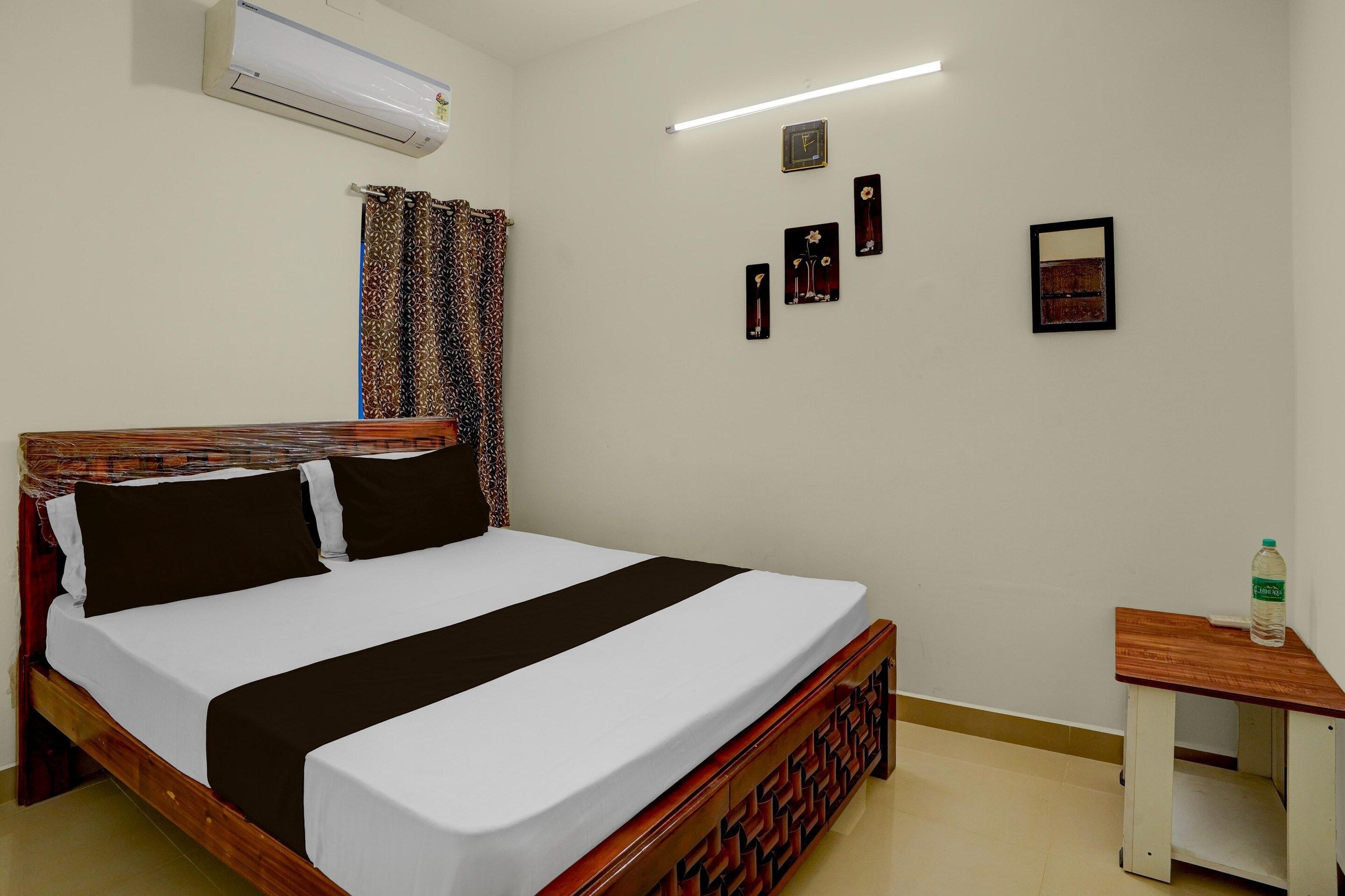 A single-bed room with a wooden headboard, a white and black bedspread, a small wooden nightstand with a bottle, and a wall-mounted air conditioner.