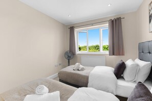 A hotel room with two beds, a window with curtains, a radiator, and a ceiling fan. - Heathrow Living 2BDR 2BA Apt 06-360stays (Stanwell)