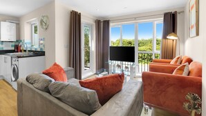 Ein modernes Wohnzimmer mit einem grauen Sofa, orangefarbenen Sesseln, einem Flachbildfernseher und einem Balkon mit Blick auf Grünflächen.