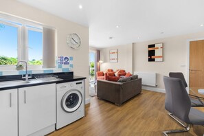 A modern kitchen with a Bosch washing machine, a dining area with a table and chairs, and a living space with a sofa. - Heathrow Living 2BDR 2BA Apt 06-360stays (Stanwell)