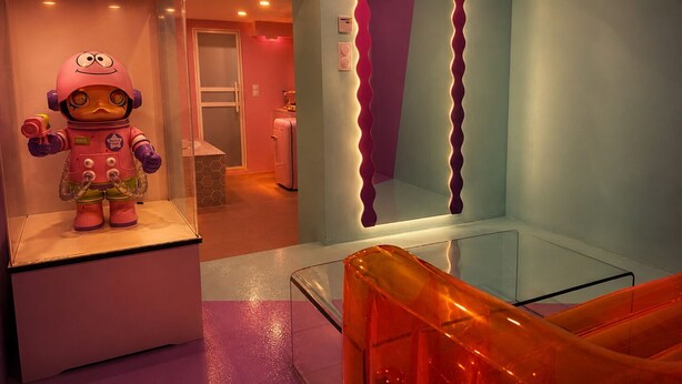 Ein Zimmer mit einer rosa Roboterfigur, einem leuchtenden Neon-Schild mit der Aufschrift 'You are Beautiful' und einer Glastrennwand.