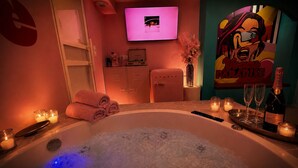 Ein Wellnessraum mit Whirlpool, Kerzen, einer Champagnerflasche und einem Fernseher an der Wand.