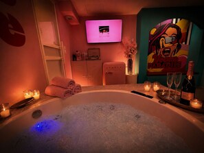 Ein Wellnessraum mit Whirlpool, Kerzen, einer Champagnerflasche und einem Fernseher an der Wand.