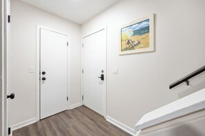 A hallway with two white doors, a wooden floor, a painting of a beach scene, and a staircase with a metal handrail. - Remodel Condo overlooks Pool walk to Celebration Park Beach & Coligny! (Hilton Head Island)