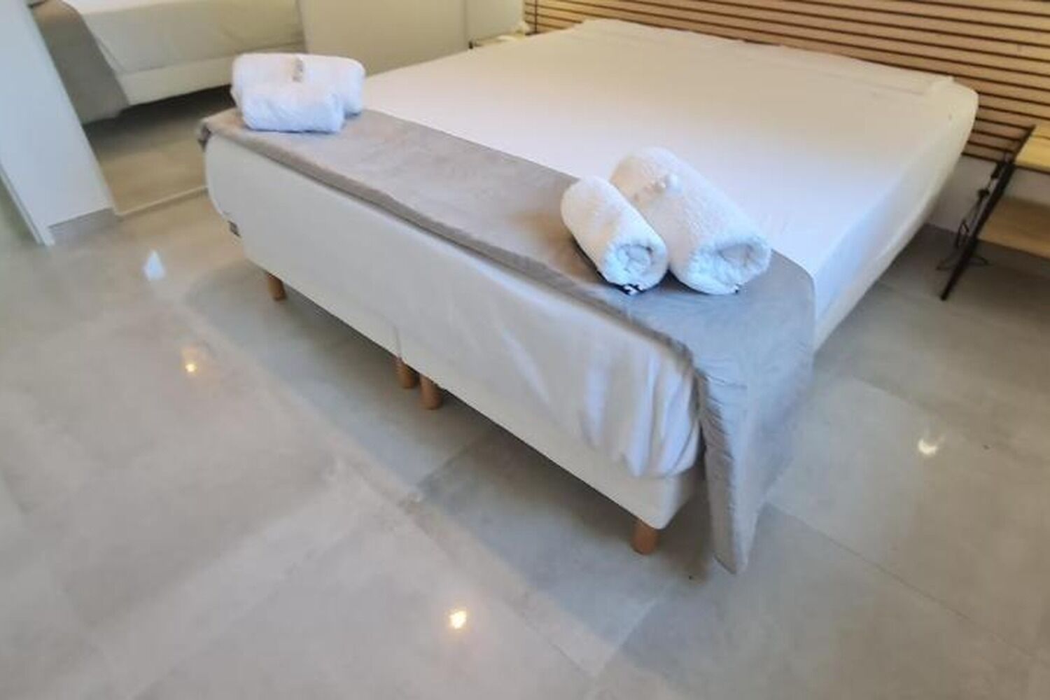 A bed with a white mattress and a grey bedspread, two rolled towels on the bed, and a white chair in the background.
