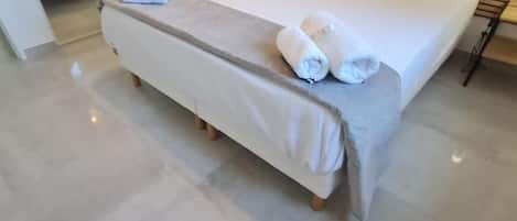 A bed with a white mattress and a grey bedspread, two rolled towels on the bed, and a white chair in the background.