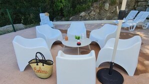 Sitzgelegenheit im Freien mit weißen Plastikstühlen und einem Tisch, einer gewebten Tasche und einem schwarzen Sonnenschirm.