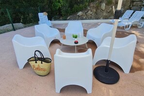 Sitzgelegenheit im Freien mit weißen Plastikstühlen und einem Tisch, einer gewebten Tasche und einem schwarzen Sonnenschirm.