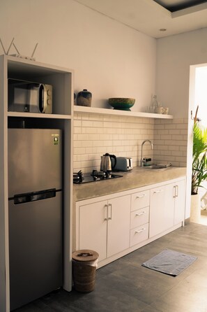 A modern kitchen with a built-in refrigerator, microwave, and stove. There is a sink with a faucet, a toaster, and a bowl on a shelf. The floor is tiled, and there is a small trash bin. - Cozy and Private 1BR Casa Su Villa in Seminyak (Badung)
